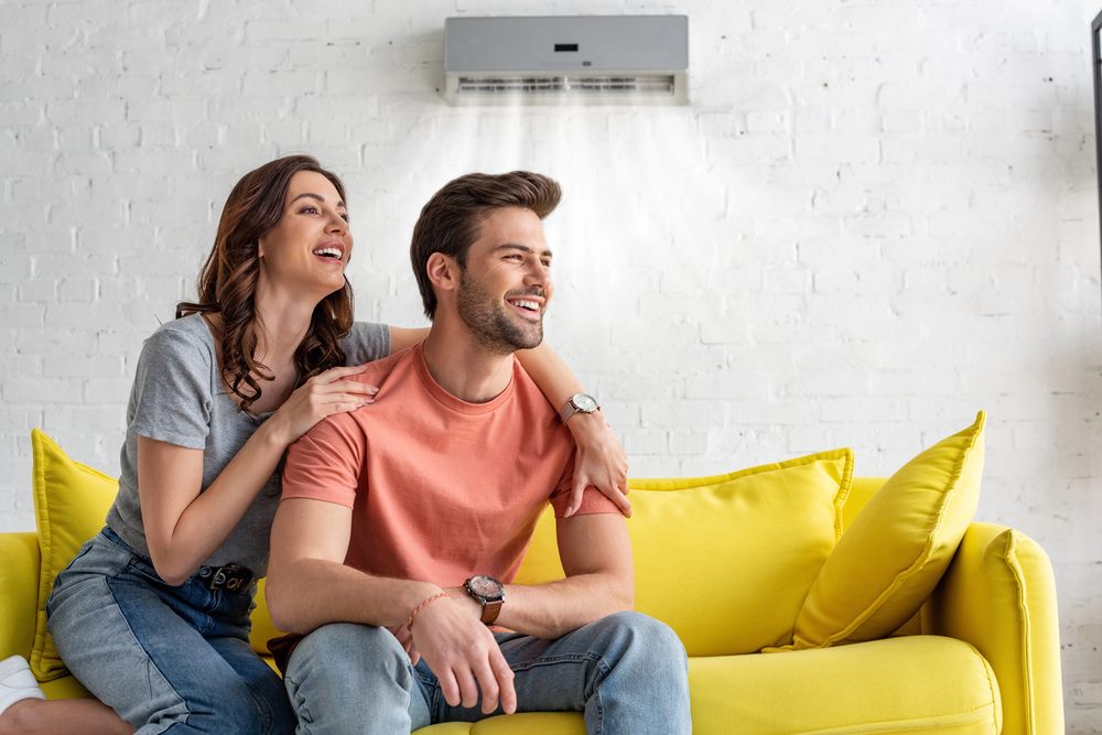 Cheerful couple sitting on a yellow sofa under an air conditioner at home, featuring Christmasair and highlighting Expert HVAC, ensuring it aligns with the page's context.