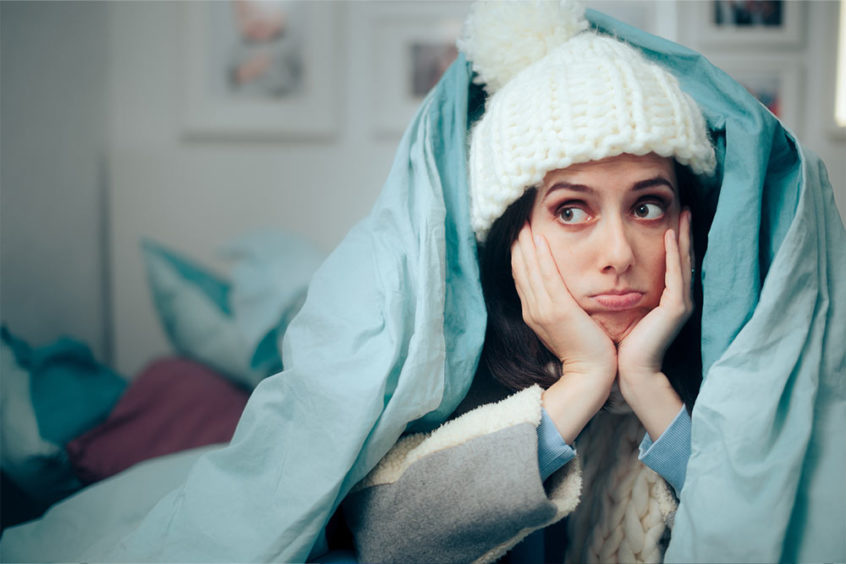 Image of a lady wrapped in a comforter, appearing chilly indoors. Featuring Christmas Air Conditioning and Plumbing, highlighting the topic “Why is my room temperature different from the thermostat setting?” to illustrate temperature inconsistencies and home comfort issues.